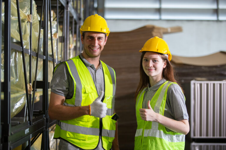 Smiling warehouse workers wearing safety gear giving thumbs up, showing teamwork, confidence, and job satisfaction in an industrial environment. Ideal for logistics and labor concepts